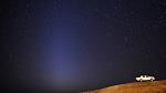 Starry night sky in the background with desert in the foreground. White truck on the side and a triangular glow of zodiacal lights in the sky.