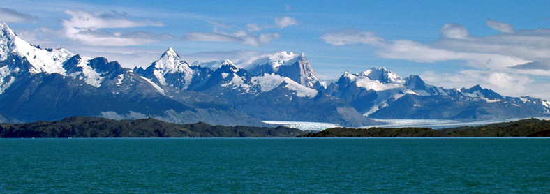 Vista de la cordillera de los Andes desde las llanuras del parque nacional Los Glaciares, la vista es desde la playa oriental de uno de los grandes valles glaciares de la Patagonia argentina, si se observa más allá del lago y de la franja boscosa y aquende a la cordillera se pueden notar a la distancia dos lenguas de glaciares que desembocan en los lagos tras bajar de las montañas siempre nevadas.