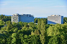 Deux bâtiments d'une dizaine d'étages dépassent d'une forêt ou d'un bois. Le ciel est bleu et l'horizon distant.