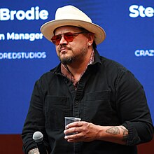 Sterlin Harjo wearing a black shirt, brown neckerchief, white Stetson hat, and red-shaded glasses, holding a plastic cup in one hand and a microphone in the other, standing onstage