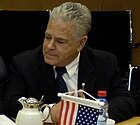 Man with grey hair and a dark suit, white shirt, dark tie sitting down, looking to his right. In front of him, a coffee pot, a bottle of water, and a small U.S. flag.