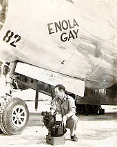 Photographie en noir et blanc d'un des membres d'équipage tenant une boîte (viseur) devant l'avion au sol.