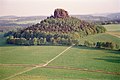 Farbfotografie in der Vogelperspektive von einer Ebene mit einem Hügel. Der Hügel besteht aus vielen Bäumen und an der Spitze ist ein brauner Felsen mit weiteren Bäumen. Die umliegende Landschaft besteht aus Wiesen, Wäldern und Hügeln.