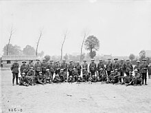 Photo en noir et blanc de militaires en tenue de combat portant des casques en deux rangées, celle de devant assise ou agenouillée et celle de derrière debout, avec des mitrailleuses