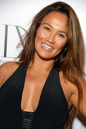 A woman with long brown hair wearing black and standing in front of a white backdrop