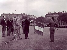 Photographie en noir et blanc montrant le président René Coty (à gauche, en queue-de-pie) entouré d'officiels, civils et militaires, qui remet la Croix de guerre des TOE au drapeau du Prytanée (brodé de la devise Honneur et discipline) que lui tend un soldat.