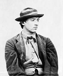 A young man looking sour and facing right. He is dressed in a jacket, bow tie, and wide-brimmed hat, and his shirt is partially unbuttoned.