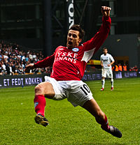 A photograph of a man dressed in a red football sweater, white sport shorts and red socks. The man is in mid play during a football match throwing himself forward for something or someone who is off-picture.