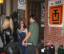 Three people mill around indoors next to a brick wall. Left to right, they include a person in a black leather jacket with long blonde hair facing away from the camera, a person in a blue dress and red sunglasses facing towards the camera, and a person facing to the left side with a green polo and dark ponytail. An orange sign points to "The Tank" on the left; another in the background reads "Collective:Unconscious".