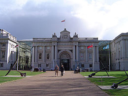 Facade of an ornate 18th century building, with tall stone columns, a wide arched entrance, balustraded roof and a central pediment from which a flag is flying. The building is approached by a wide path flanked by lawns.