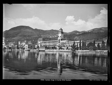 Schwarz-Weiß Fotografie, auf der man eine Häuserzeile sieht, die sich im Wasser eines Sees spiegelt. Im Hintergrund bewaldete Hügel.