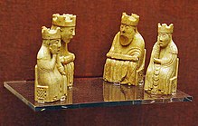 Four gold-coloured chess pieces sit on a glass shelf with a red wall in the background. All four pieces are seated and wear crowns. Two of the pieces are larger and have beards, the other two are female figures.