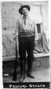 A black-and-white photograph of a man in a cowboy hat, a cartridge belt around his waist and a rifle by his foot, looking straight at the camera