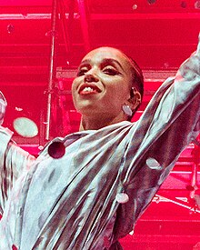 FKA twigs, against a red-lit background, is smiling to the crowd.