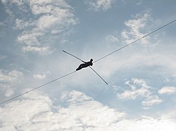 A small figure holds a balancing pole while lying on a high-wire