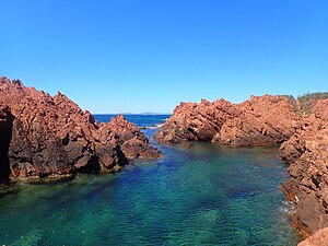 Cliché numérique couleur. Sur un ciel bleu se détachent des roches rouges plongeant dans la mer.