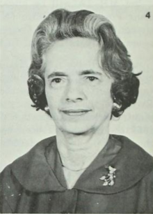 An older white woman with set grey hair, wearing a round-collared dark jacket with a brooch