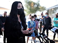 Cher, wearing a black face mask and long dark hair, stands at an outdoor event with microphones in front of her and a crowd of masked people in the background.