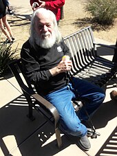 An elderly Joe Proski sitting down on a bench while attending a funeral service.