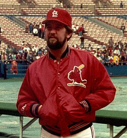 A bearded man with his hands in his jacket pocets, wearing a red baseball cap and red jacket with a bird logo on the left chest, standing with a field and stands in the background.