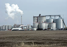 Usine de distillation en arrière plan, avec de grands silos à droite et une cheminée rejetant une fumée blanche à gauche, et un champ avec la terre laissée à nue au premier plan.