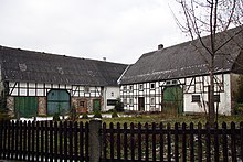 Hinter einem Gartenzaun: rechts Fachwerkbau mit grünem Deelentor, rechter Gebäudeteil vermutlich Stallungen, links Wohnbereich, vor dem Haus eine Bank. Im rechten Winkel dazu Verbindungsbau und weiterer Fachwerkbau, niedriger. Ebenfalls mit Deelentor (grau), unterer Gebäudeteil aus Bruchstein. Beide Häuser sind schwarz eingedeckt.