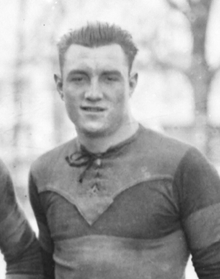 Photographie noir et blanc : c'est un portait en buste d'un homme en maillot de sport qui regarde devant lui. L'arrière-plan est flou et on devine des arbres.