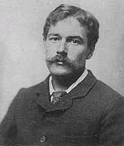 A bust-length monochrome photograph of a white male with a handlebar moustache; wearing a jacket and tie.