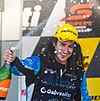 A male in his mid-20s in racing overalls sporting long hair with a yellow sponsored baseball cap on his head celebrating with a bottle of champagne in his right hand