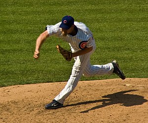 A man standing on a brown mound, with green grass surrounding, is throwing towards the left. He is wearing a white shirt, white pants, and a blue hat.