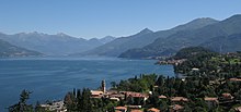 Panorama d'un lac de couleur bleu azur, bordé par les montagnes. On distingue une ville dans la forêt qui borde le lac.