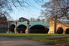 A bridge with three arches spanning a wide river. The near bank is grass with some trees bare of leaves. The sky is blue with a couple small white clouds.