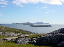 A rocky moorland in the foreground and low, dark islands against a blue sky in the background.