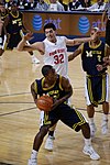 A basketball player in a dark blue uniform is maneuvering with a basketball while the defender in white is in the air behind his back.