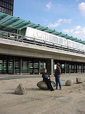 Two people look up at a train stopped at an elevated glass and concrete station