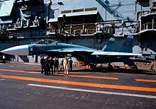 Aqua and blue jet aircraft on aircraft carrier deck, with a group of men standing close by. Behind the jet is the ship's island