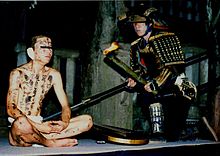 A man in black armor kneels beside a man clad only in a loincloth. The second man has black symbols painted on his body and bandages over his ears.