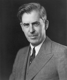 Black-and-white image of the head and shoulders of man about fifty with upswept hair, wearing a gray suit and a dark tie