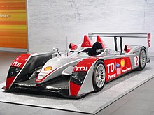 A front-three quarters view of the Audi R10 that won the 2007 12 Hours of Sebring. The car has the number 2 and the words "TDI Power" emblazoned on the sides, and just above the rollover hoops are the drivers' names, Emanuele Pirro, Marco Werner, and Frank Biela. The car is finished largely in gloss silver, the wheel arches and sidepods are finished with a bright red as are the rollover hoops above the drivers' head, and the rear wing and wheels are both a brilliant silver