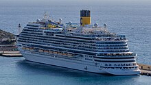 The port-side and aft of a cruise ship with white paint, many decks of balconies, and a yellow vertical cylindrical funnel with a blue 'C' painted on it, is shown from a distance.