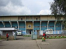 Vue extérieure de l'entrée du stade. Un portail ouvert figure au premier plan.