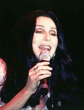 Cher sings into a microphone, her long dark hair framing her face, wearing a simple, casual light-colored outfit and a ring.