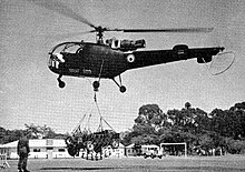 A green helicopter marked with a blue-white-red roundel; the roundel of the Federal Rhodesian Air Force. Against a backdrop of observers and barracks, it is lifting a Rhodesian Army jeep via a cargo net.