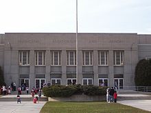 The entrance of a concrete building behind a front lawn and a flag pole