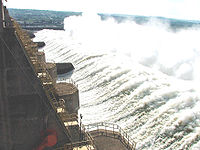 Aerial view of a concrete structure, a low hill and massive amounts of water that spur in a waterfall like way from the structure