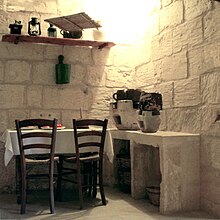 La photographie couleur montre une cuisine en pierre blanchie à la chaux. Une table couverte d'un nappe blanche et deux chaises occupent la moitié gauche. La droite est meublée d'un plan de travail portant des braseros de cuisson. En dessous, des paniers sont rangés. Au-dessus de la table, une étagère porte des lampes anciennes, un moulin à café et un vase.