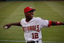 With baseball in hand, an African-American man wearing a white and red Nationals baseball uniform cocks his arm backward as he prepares to throw