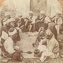 An old photo of a dozen old and middle-aged men sitting on a ground around a mat. A man in front sits next to a mortar and holds a bat, ready for grinding. A man opposite to him holds a long spoon.