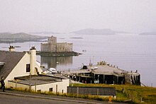 A white building sits above a concrete pier. Offshore there is a small island on which there is a building with grey featureless walls. Various other islets can be seen in the background through the mist.