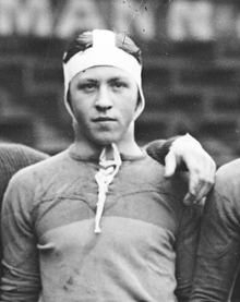 Photographie noir et blanc : c'est un portait en buste d'un homme en maillot de sport avec bonnet qui regarde devant lui. L'avant-bras de quelqu'un est posé sur son épaule gauche. L'arrière-plan est flou et ressemble à un mur de brique sur lequel on distingue les lettres "MANN".